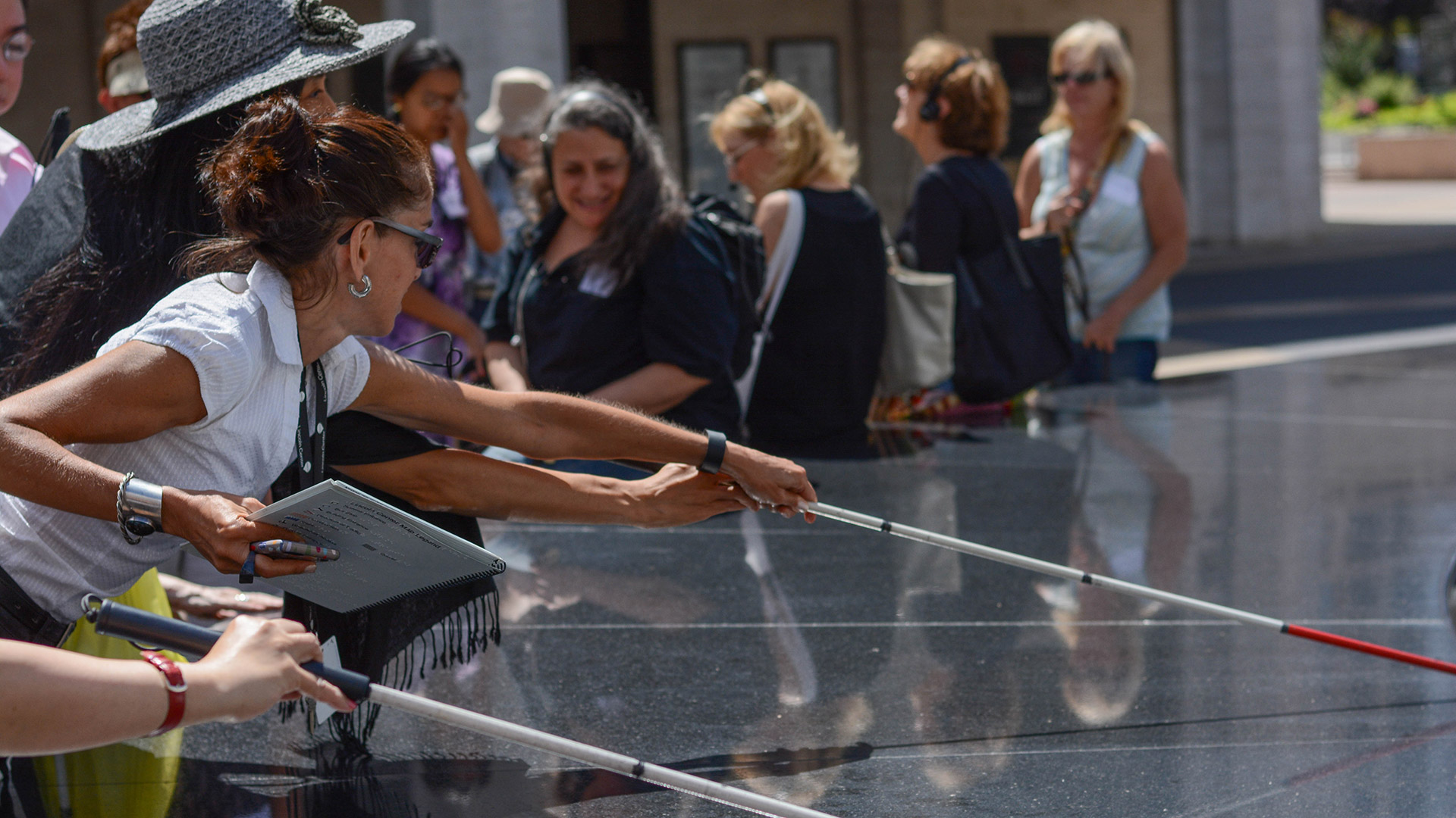 A group on a touch tour of Lincoln Center. A woman reaches out over the black granite rim of the Revson Fountain using a cane. Others on the tour watch and smile.